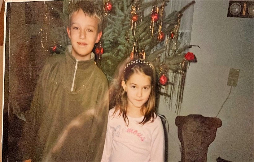 Vor einem geschmückten Weihnachtsbaum stehen ein kleiner Junge und neben ihm ein kleines Mädchen.