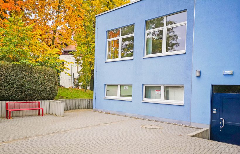 Die Schule der DPFA ist ein hellblaues Gebäude umringt von viel grünen Bäumen auf dem Schulhof.