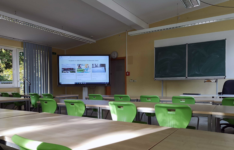Grüne Stühle stehen in einem Klassenraum. Vorn neben der Magnettafel steht eine digitale Tafel.